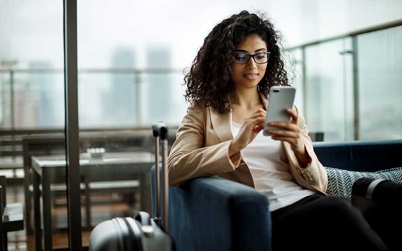 Woman sitting in an airport departures lounge on her phone with suitcase next to her