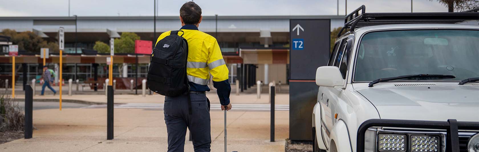 Man wearing hi-vis with a back pack and suitcase walking towards Perth Airport Terminal 2